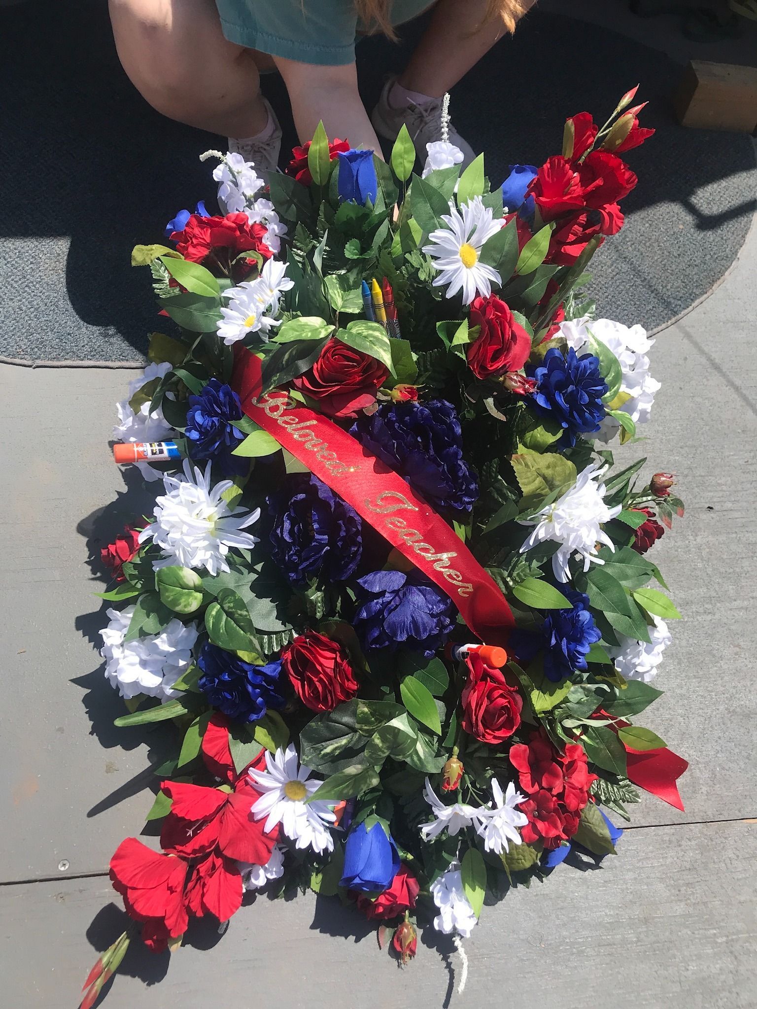 Red, white, and blue floral wreath with a red ribbon, displayed outdoors on pavement.