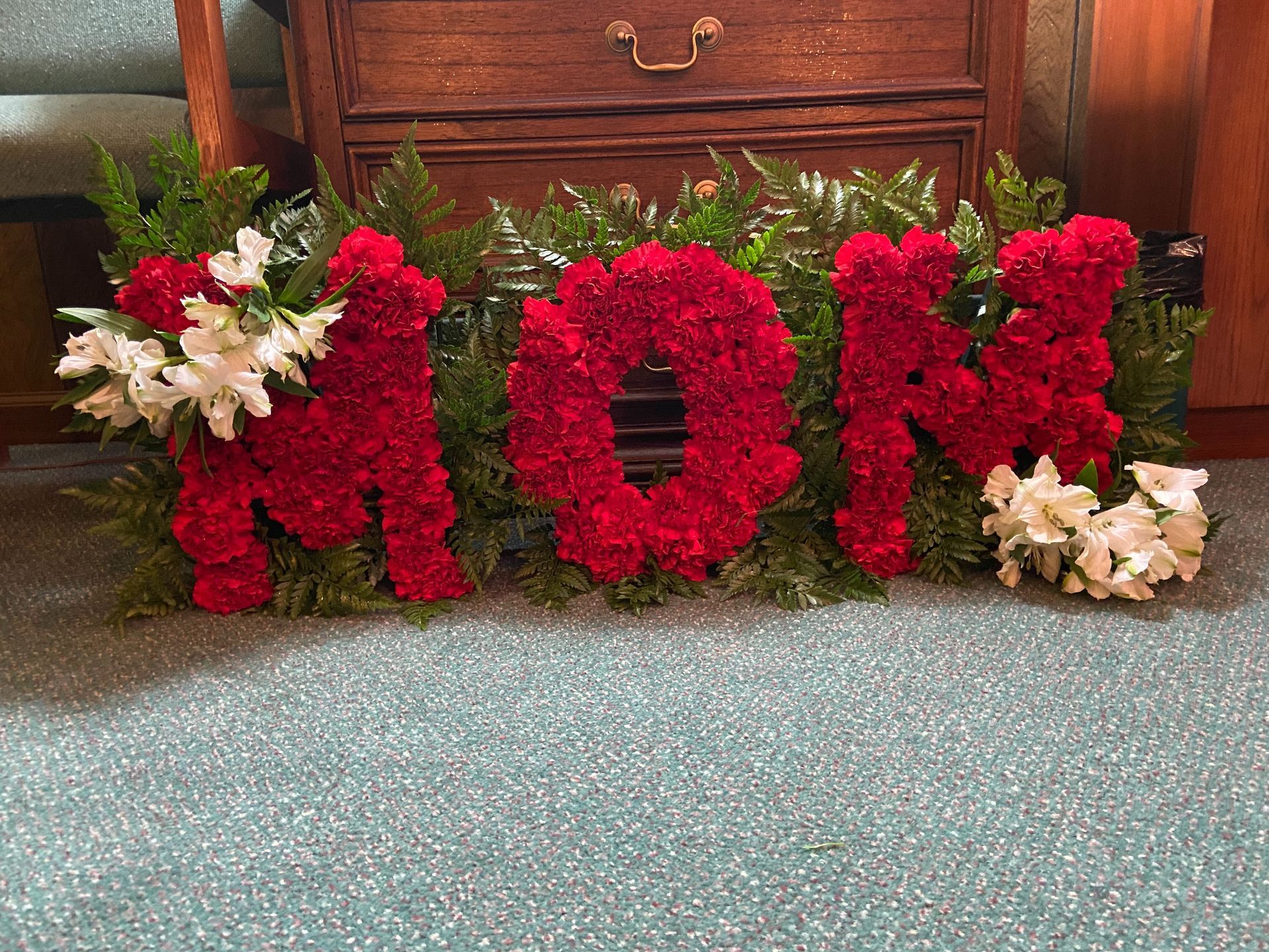 Red floral “MOM” decoration with white flowers, set on a carpeted floor in front of furniture.