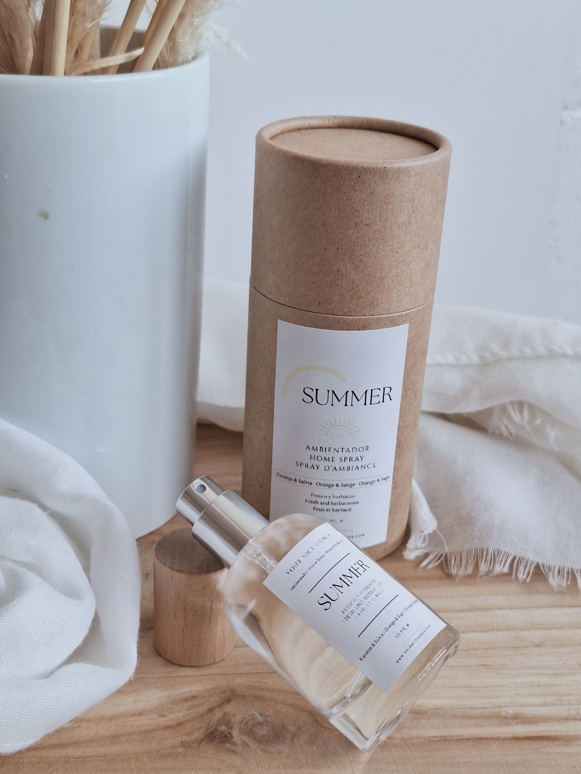 Een doorzichtige glazen parfumfles met een zilverkleurige spraydop naast een houten blok op een witte stoffen ondergrond.