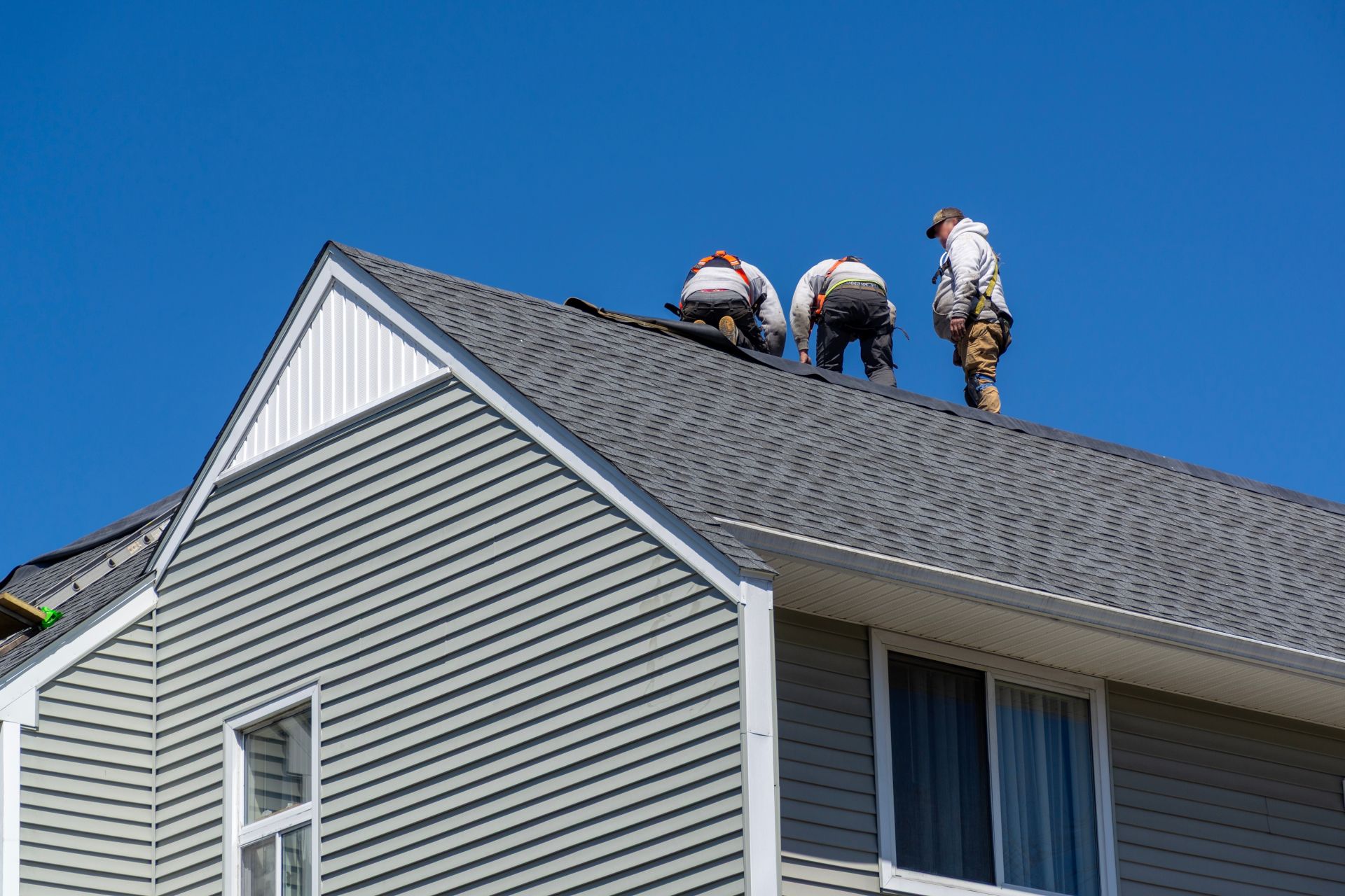 Workers repairing the roof of a gray house under a clear blue sky