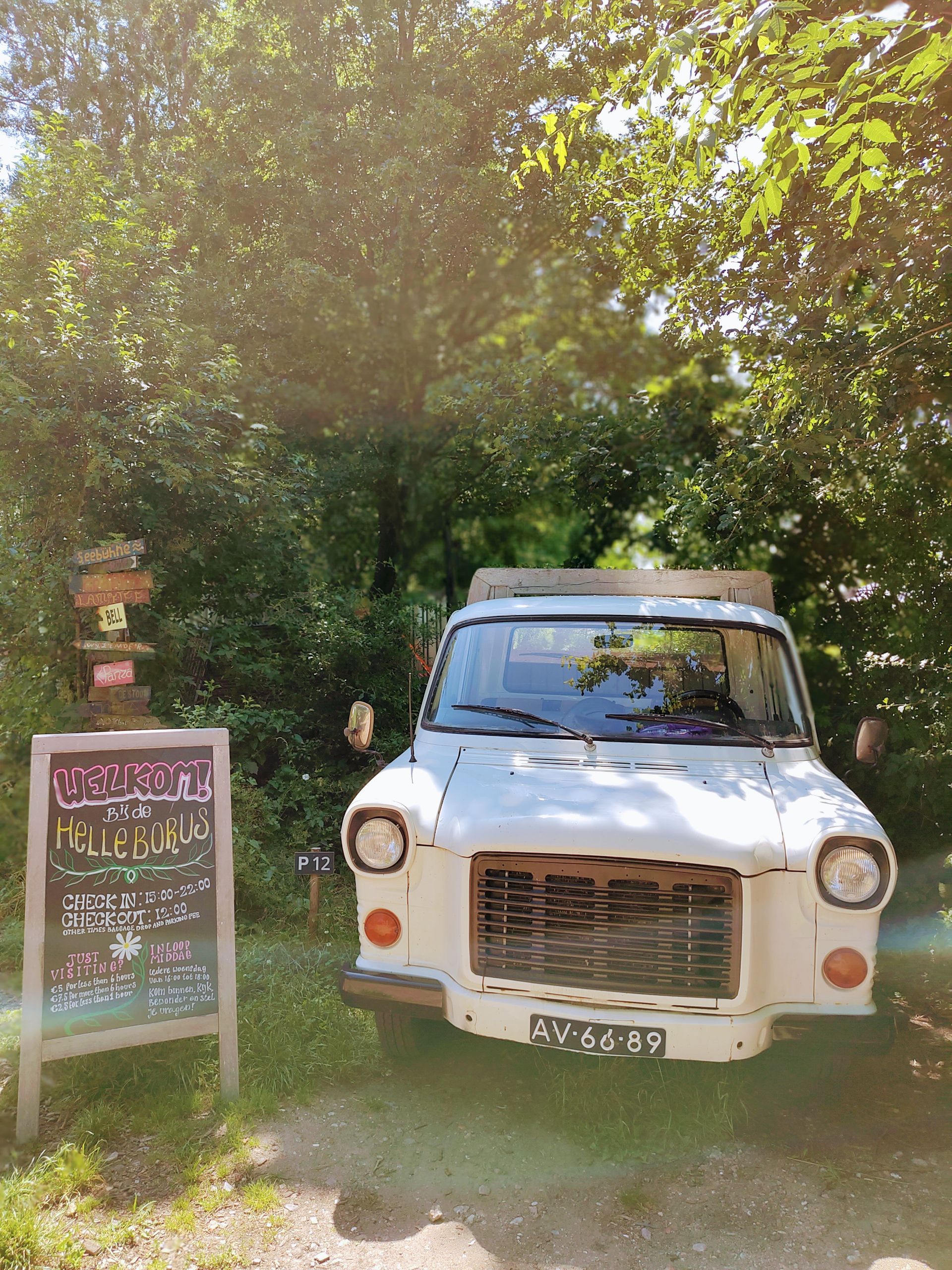 Een witte oldtimertruck staat overdag geparkeerd naast een krijtbord, voor weelderige groene bomen.