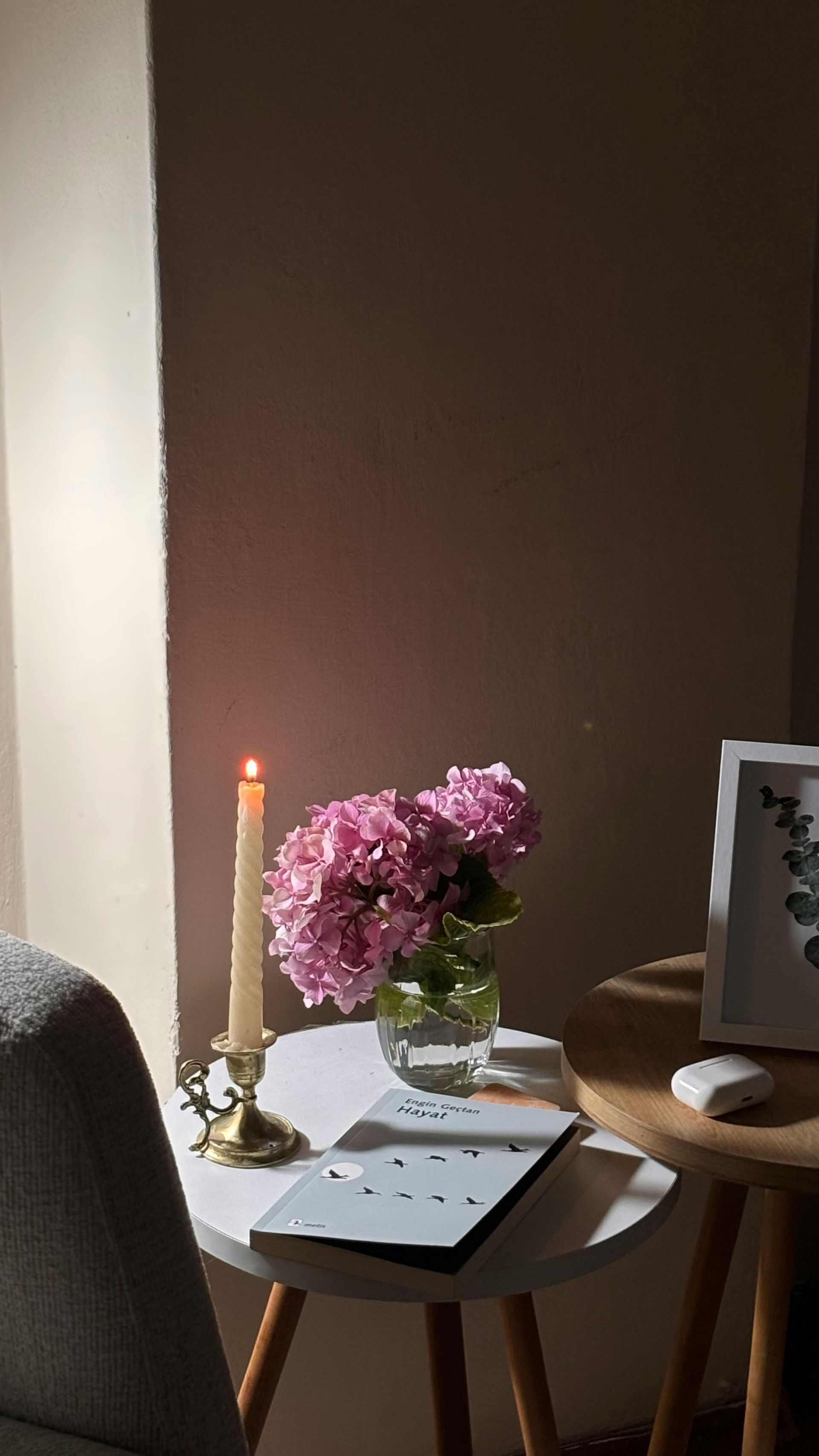Een scène bij kaarslicht: roze bloemen, een brandende kaars en een open boek op een tafeltje bij een raam.