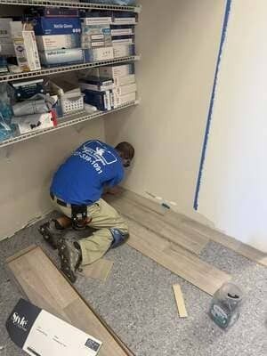 Worker kneeling on a floor, installing baseboard in a room with shelves of supplies.