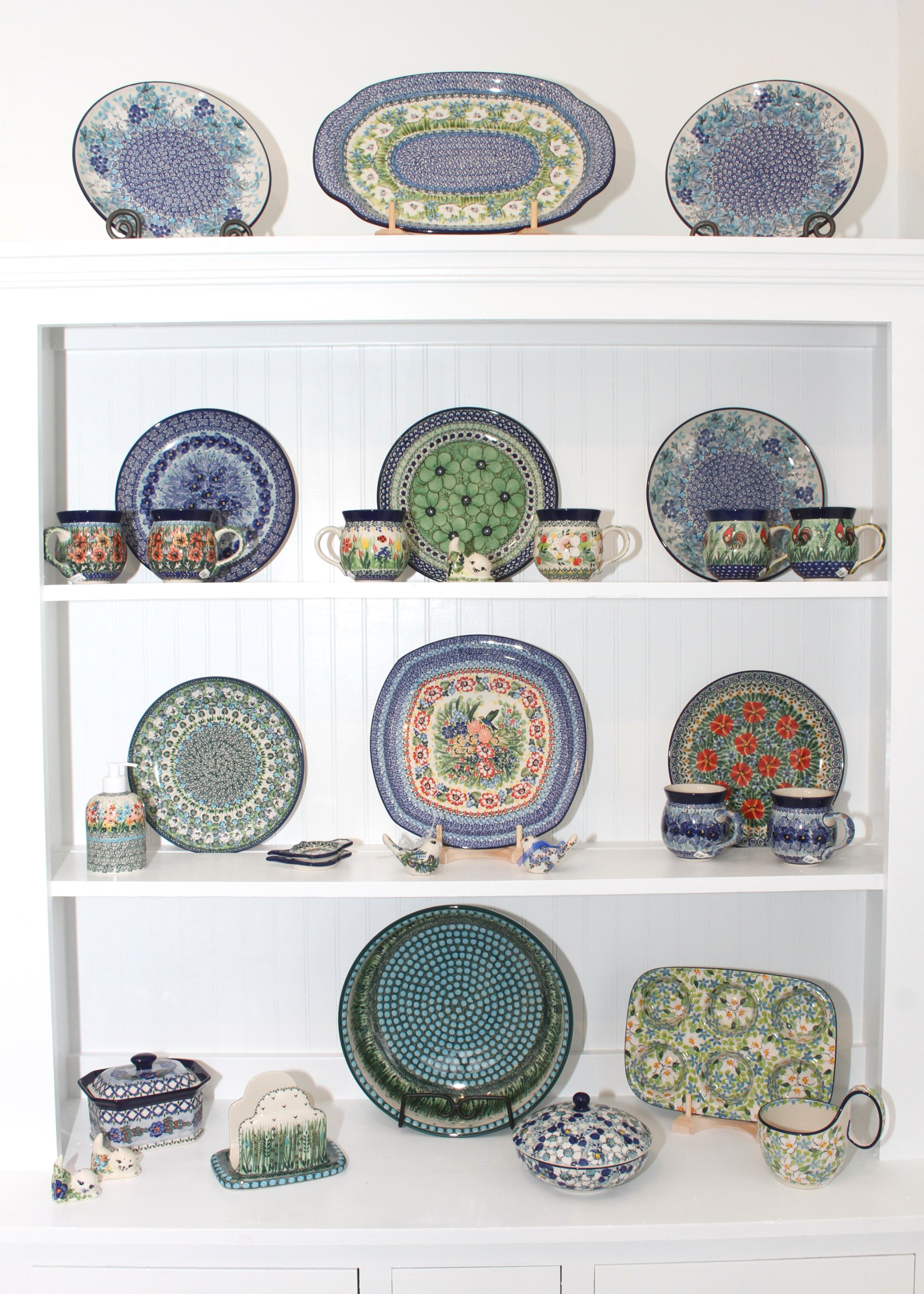 White cabinet displaying Polish pottery in blue, green, and floral patterns.