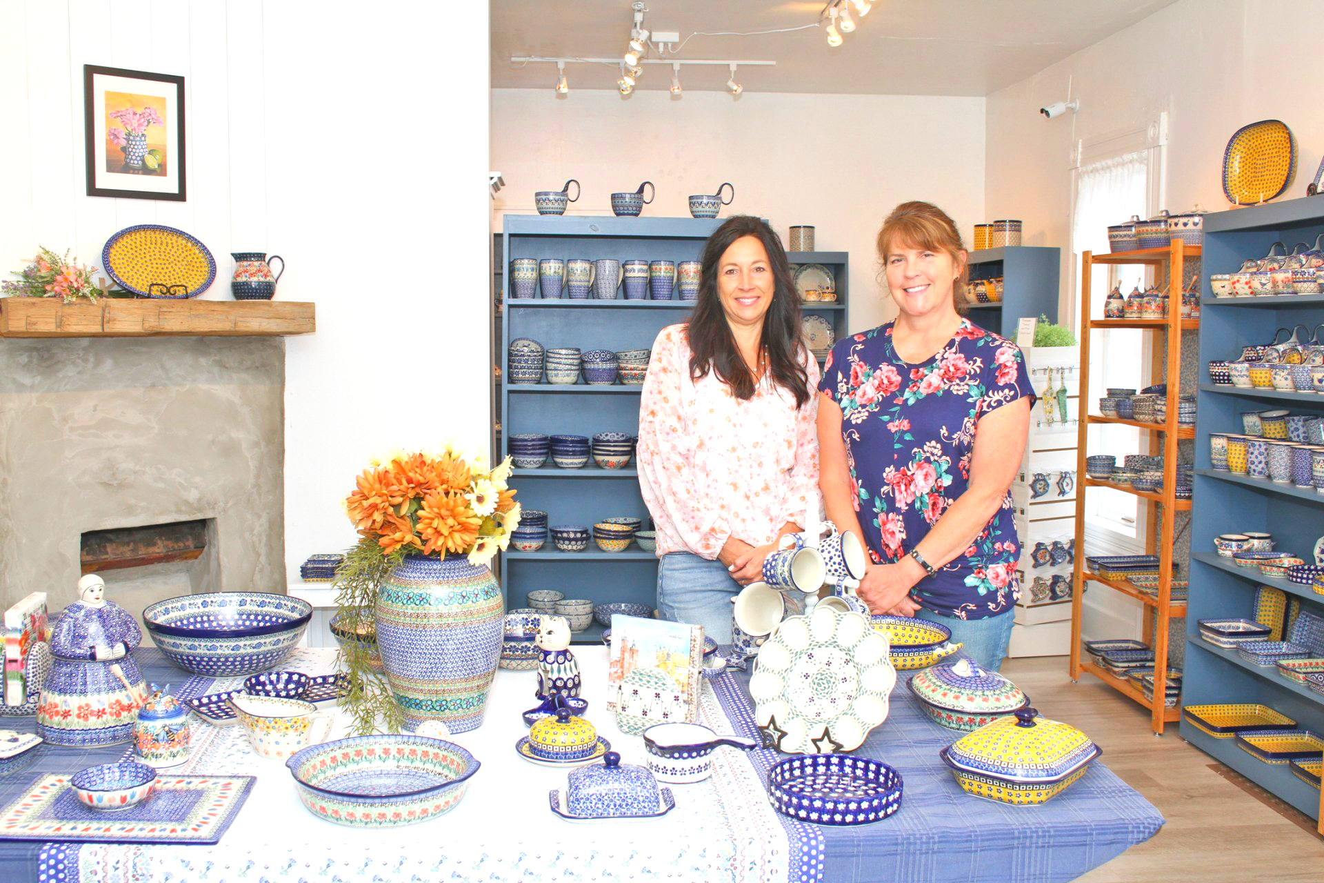 Two women in a shop filled with blue and white pottery. They stand behind a table of items.
