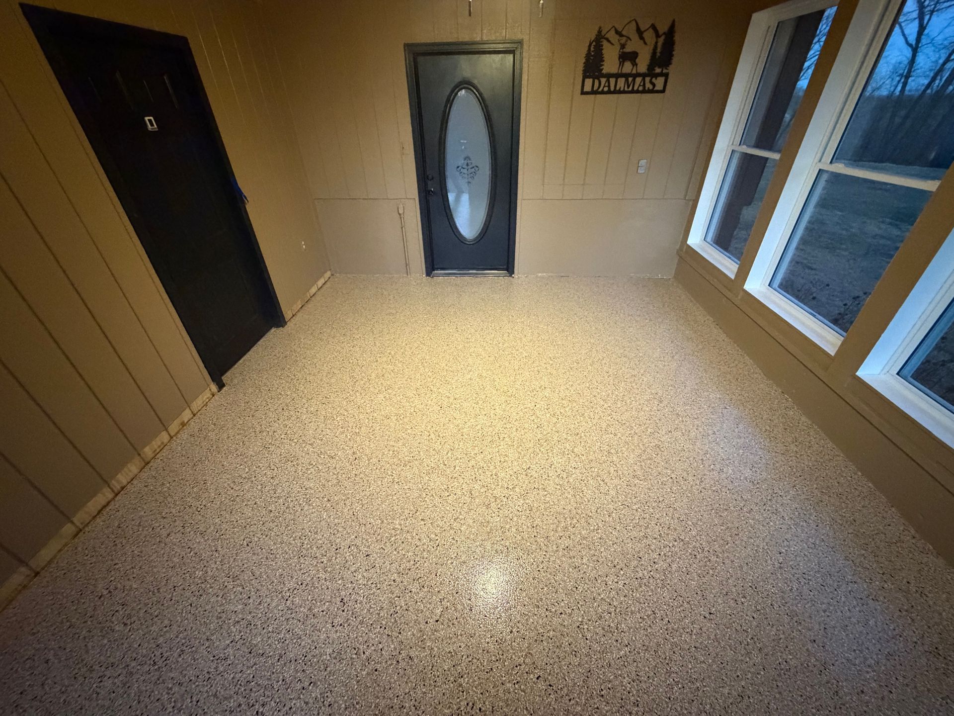 Empty beige room with terrazzo floor, black door, oval-glass door, wall sign, and windows on the right.