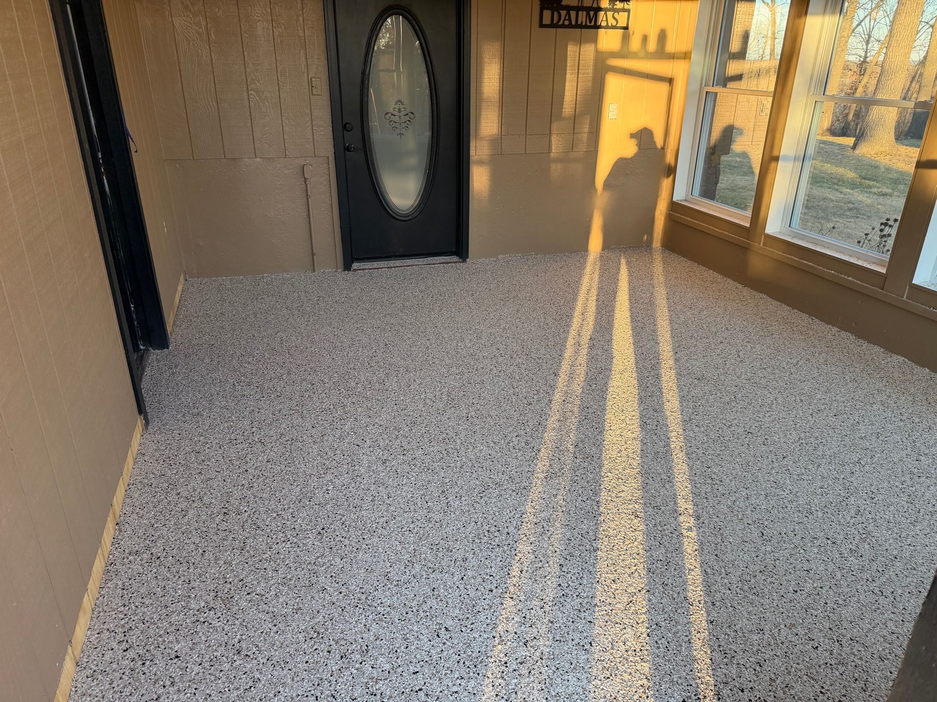 Sunlit apartment balcony with speckled carpet, black door, and windows casting long shadows.