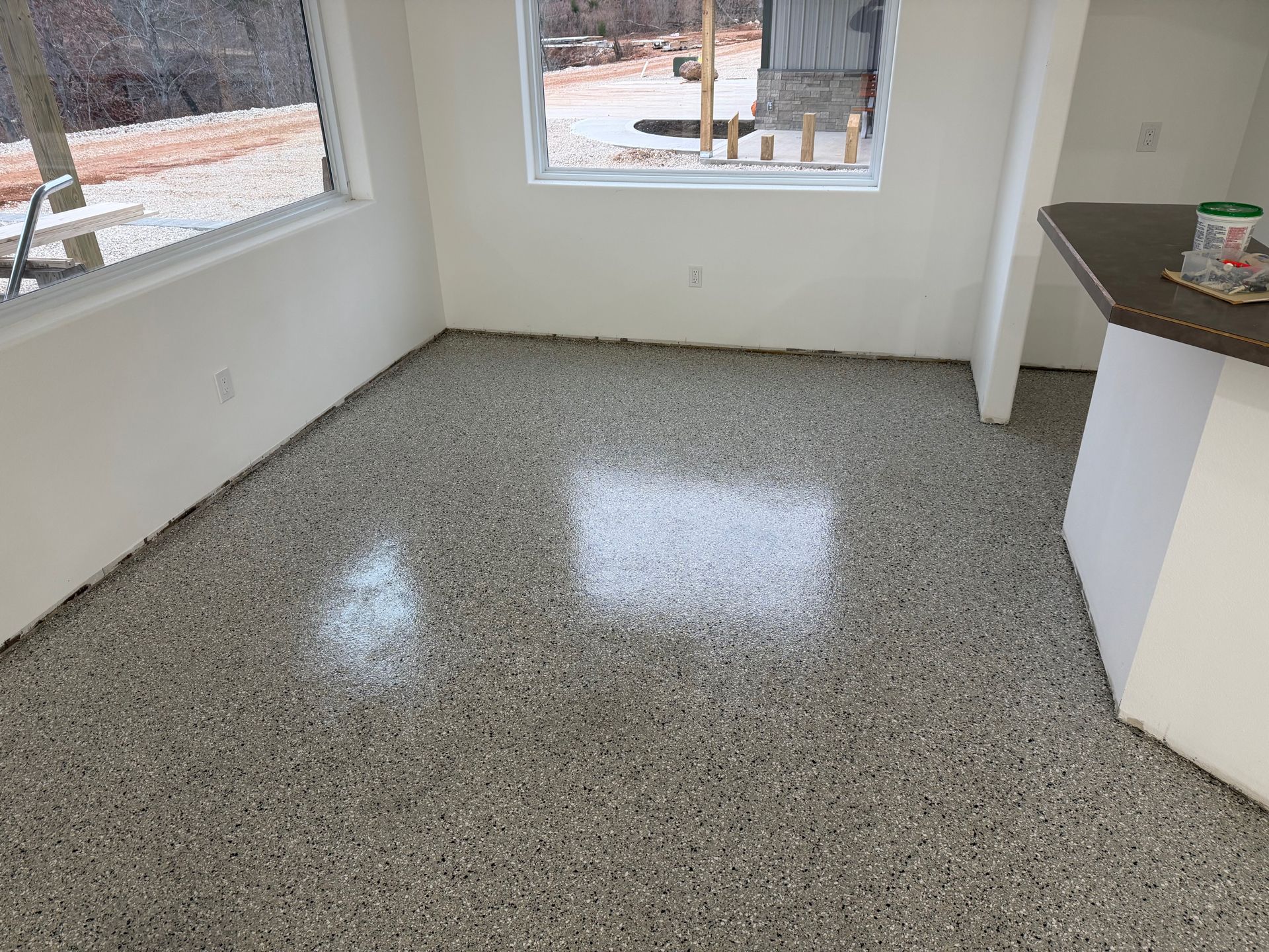 Empty sunlit room with speckled gray floor and large windows.