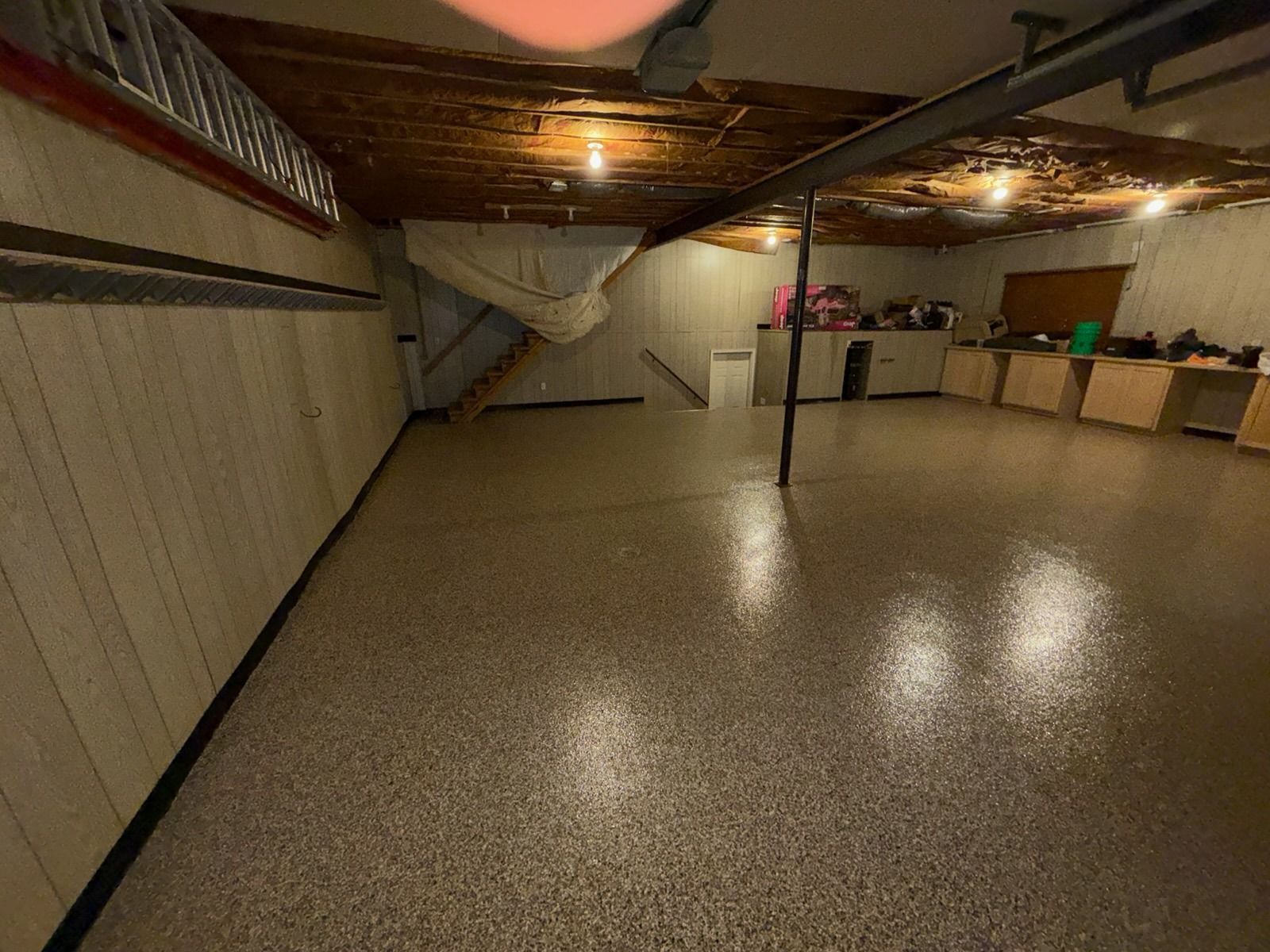 Basement room with speckled epoxy floor, exposed ceiling beams, and a long storage counter.