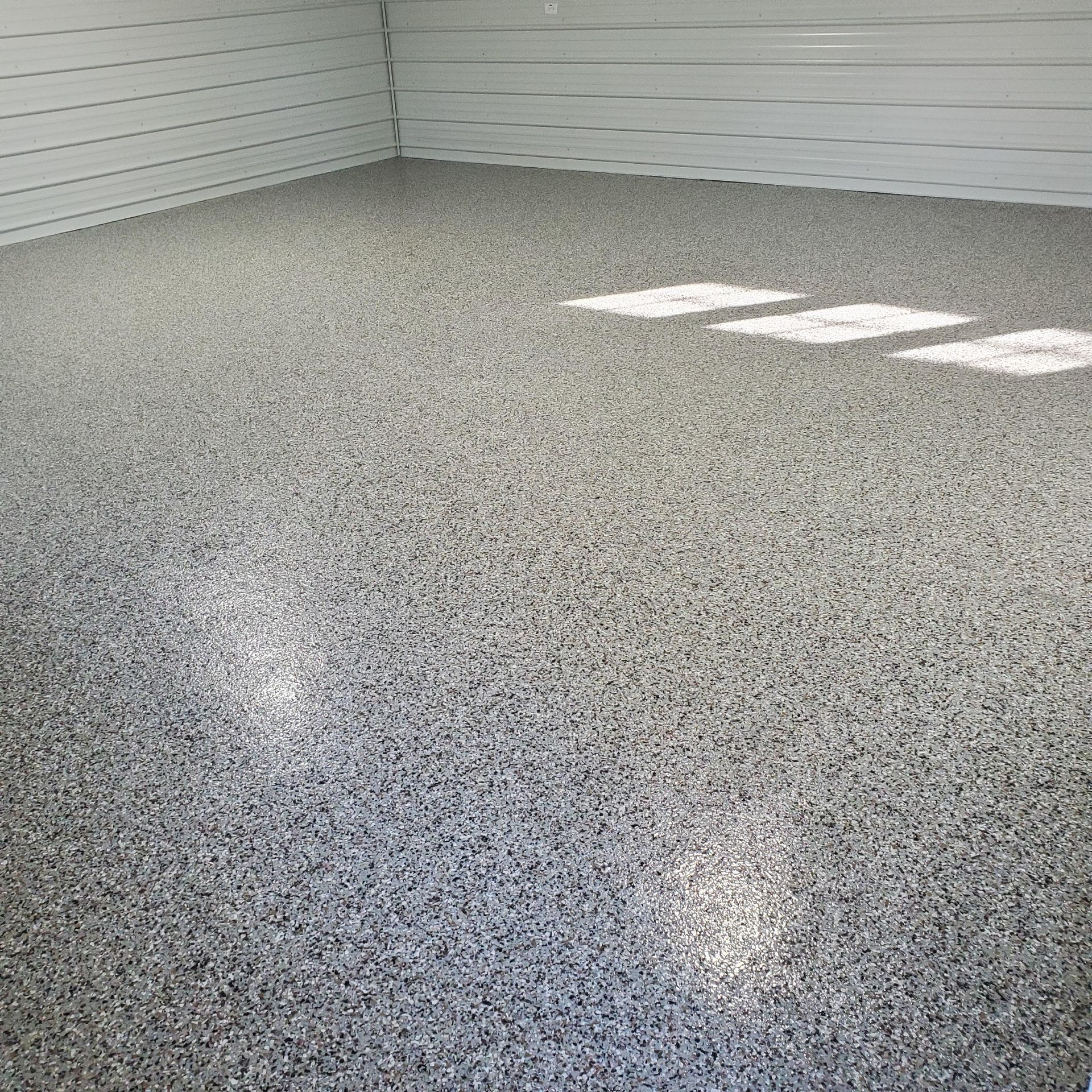 Empty room with gray speckled epoxy floor and white paneled walls, lit by bright window reflections.
