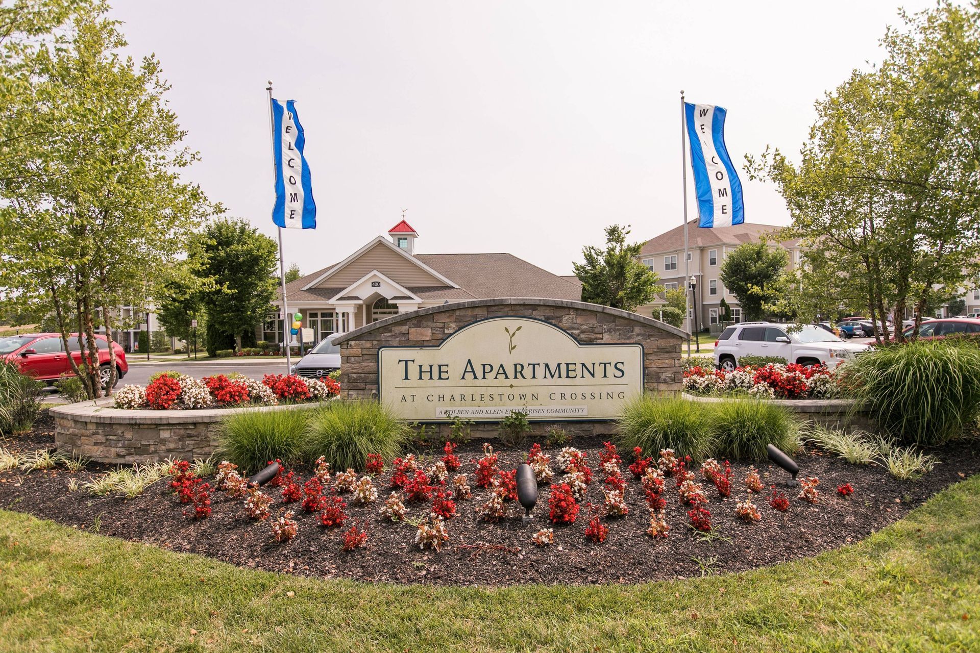 The Apartments sign with flower beds and blue flags in front of a building entrance at Apartments at Charlestown Crossing.