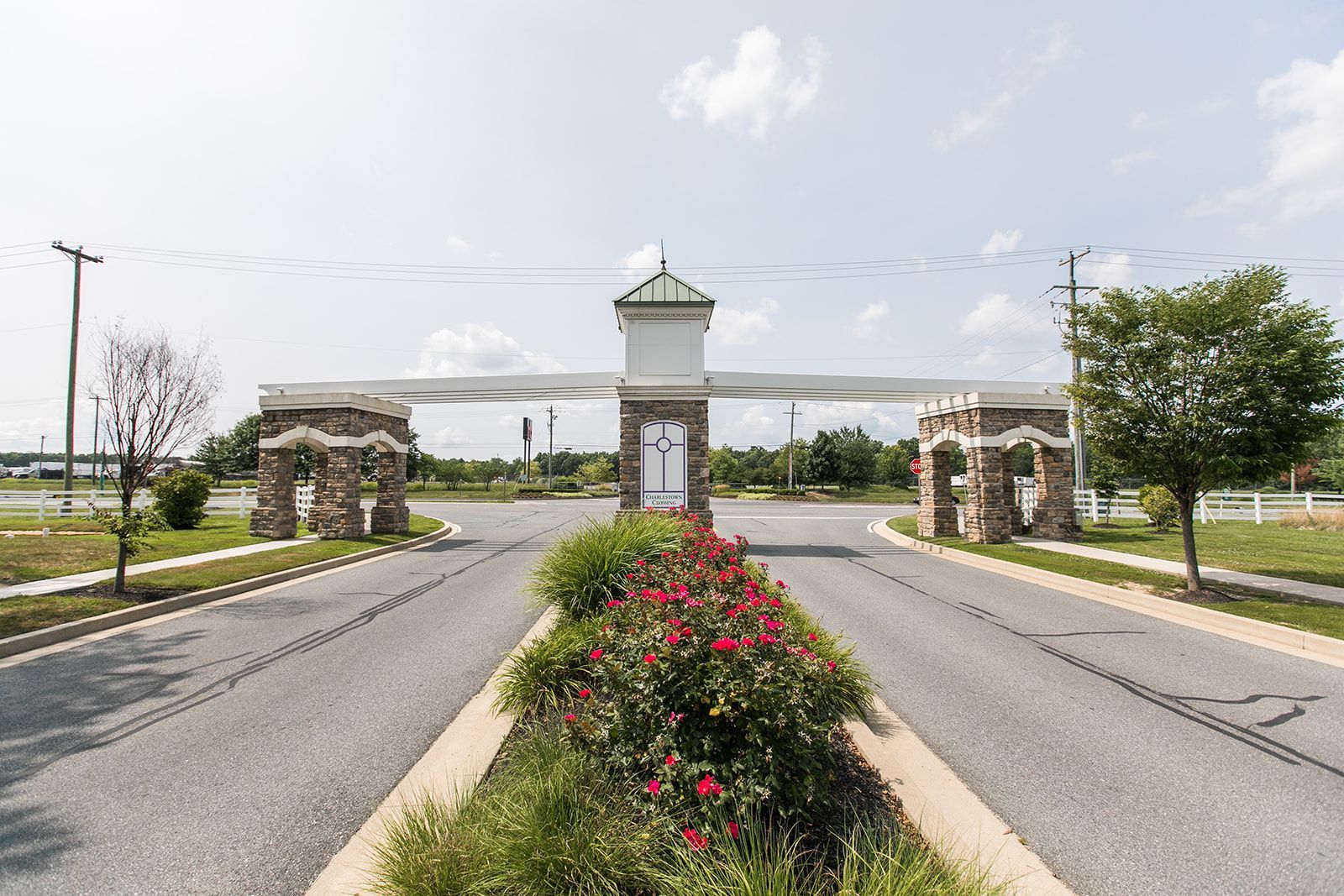 Gated entrance with stone pillars, central tower, and flower-lined road under a bright sky at Apartments at Charlestown Crossing.