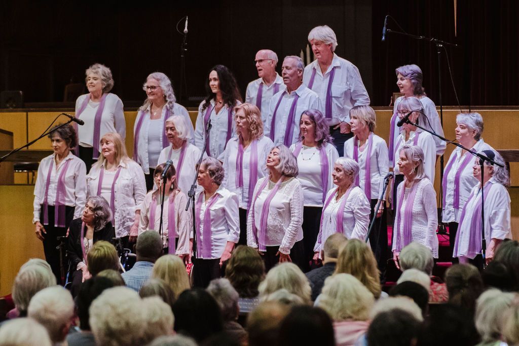 Choir in white and pink outfits standing on a stage in a concert hall, with an audience in front
