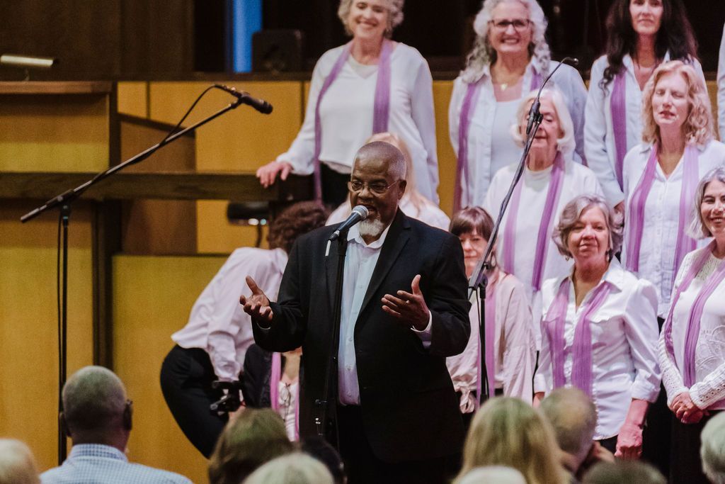 Choir conductor leading a group of singers on a stage, with piano and microphone in the background.