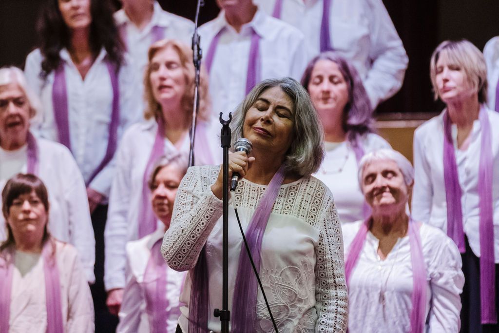 Singer performing into a microphone in front of a choir wearing white and purple scarves