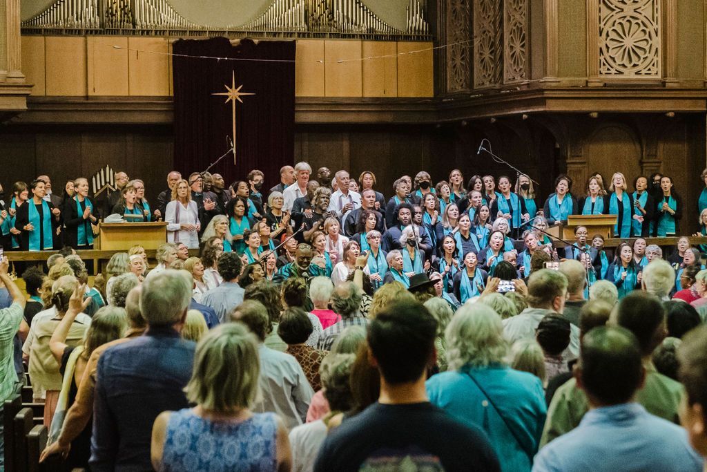 Choir performing on a church stage before a standing audience in a wood-paneled hall