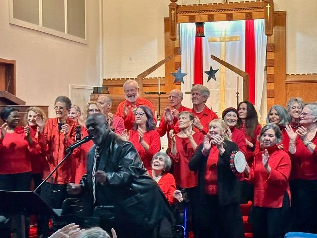Choir in red jackets singing on a church stage with a piano in the foreground