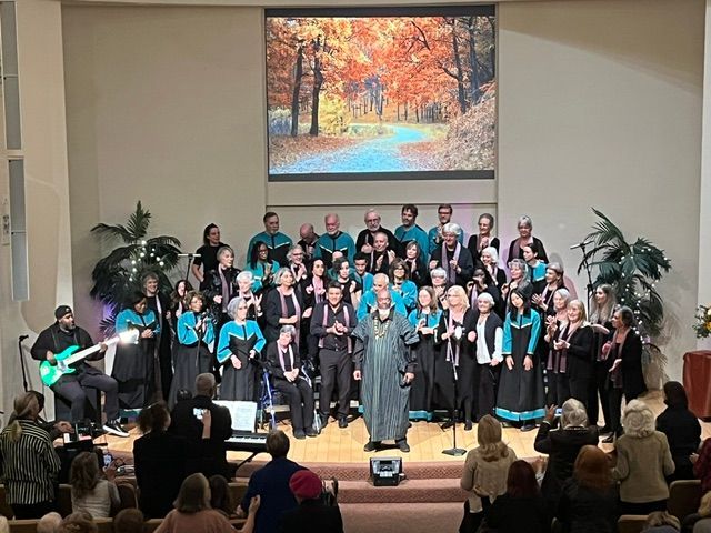 Choir performing on church stage with autumn backdrop, conductor centered, audience seated in front