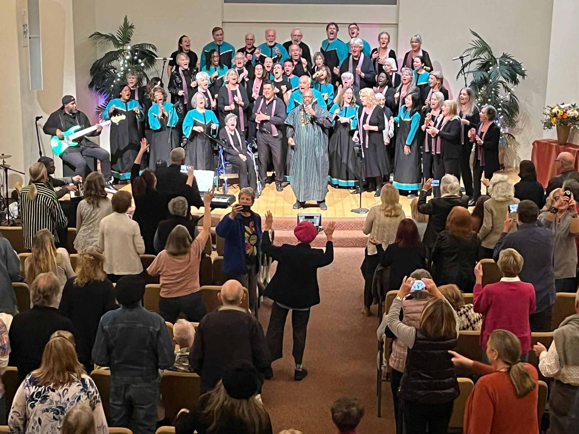 Choir performing on stage in a church, with audience standing and clapping in the foreground.