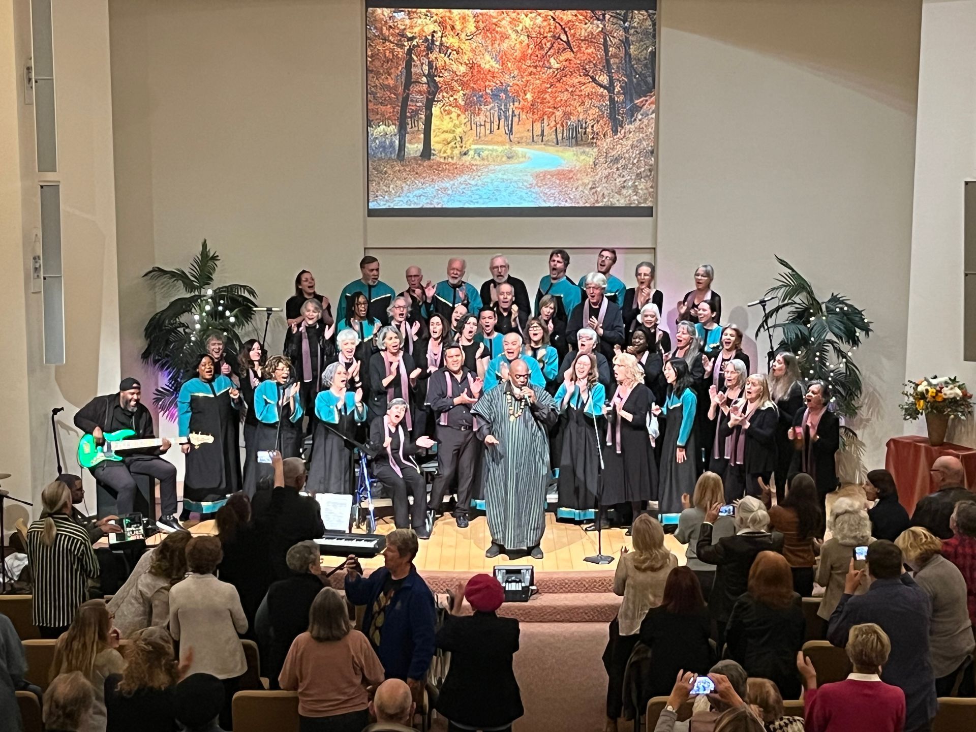 Choir on church stage in black and teal, with audience seated below and autumn landscape painting overhead