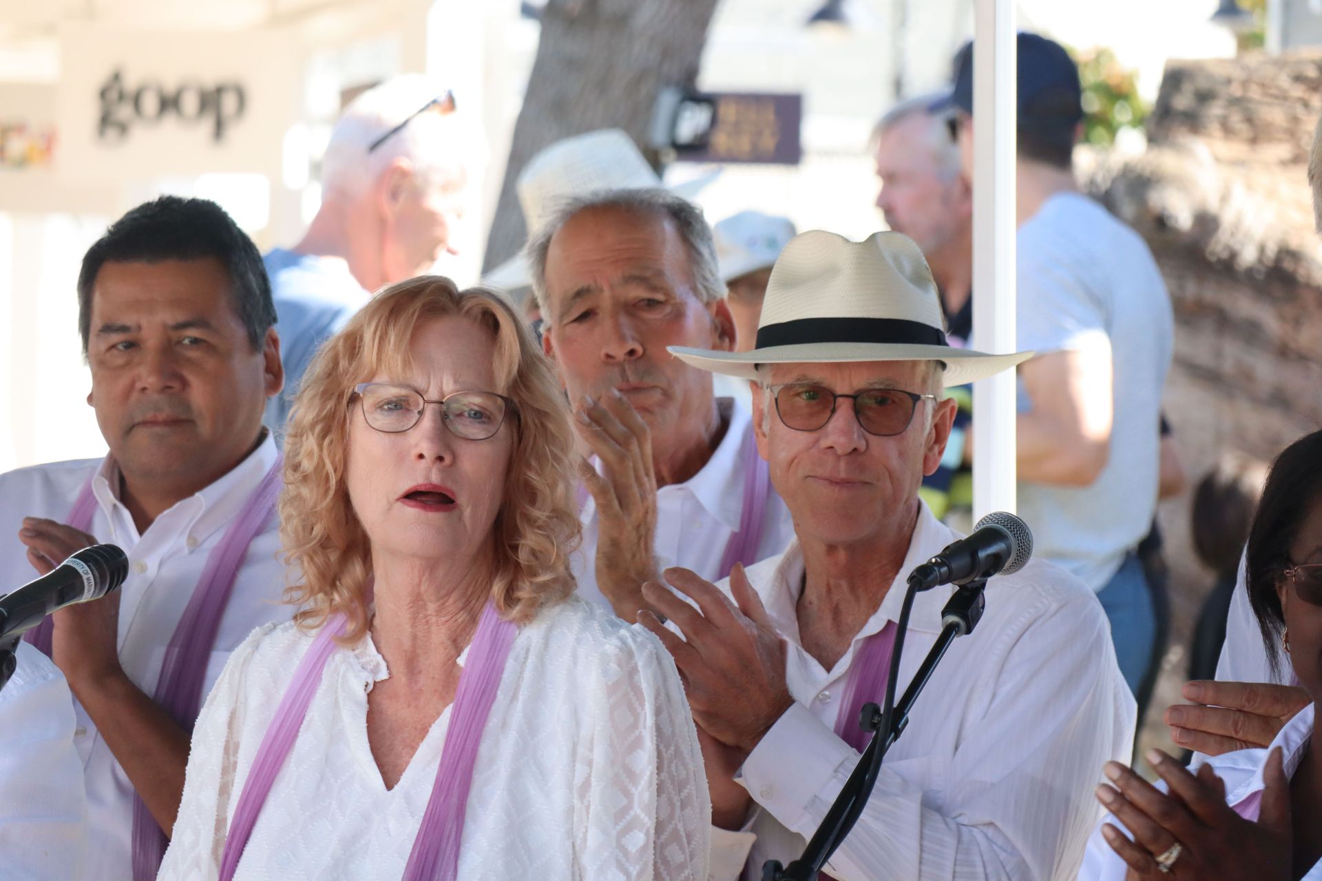 People speaking at microphones at a public outdoor event, with a woman in white in front