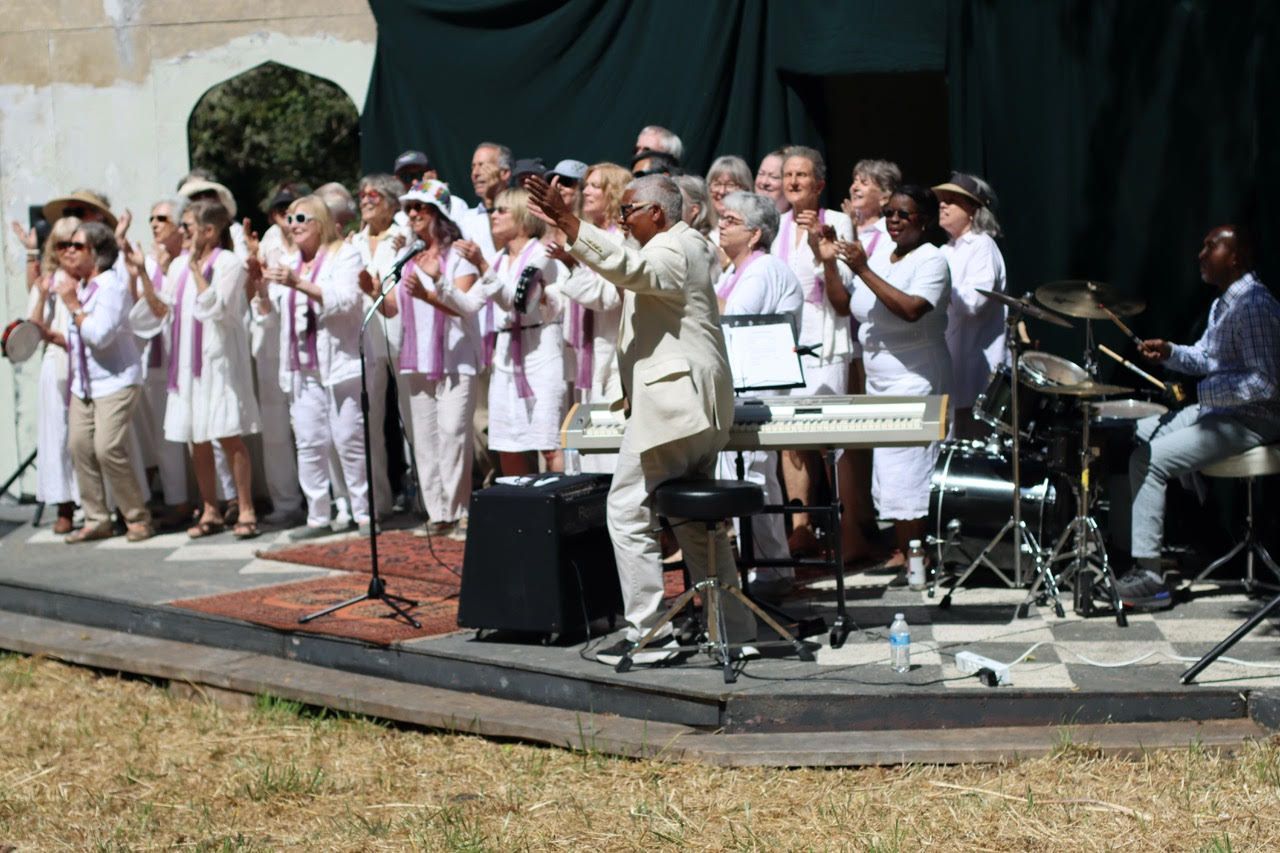 Children singing on an outdoor stage, with a conductor, microphones, keyboard, and drum kit