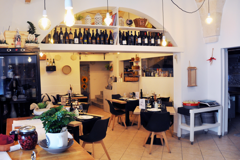 Accogliente interno da caffetteria con tavoli in legno, sedie nere, lampade a sospensione e scaffali con bottiglie e oggetti decorativi.
