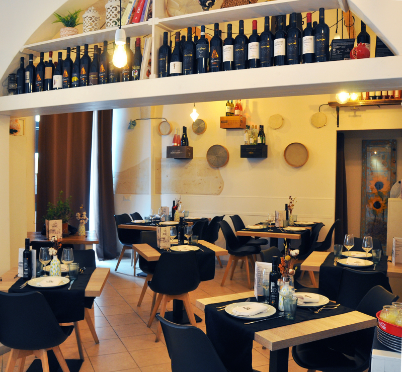 Accogliente sala da pranzo di un ristorante con tavoli apparecchiati, sedie nere, decorazioni alle pareti e bottiglie di vino esposte in alto.