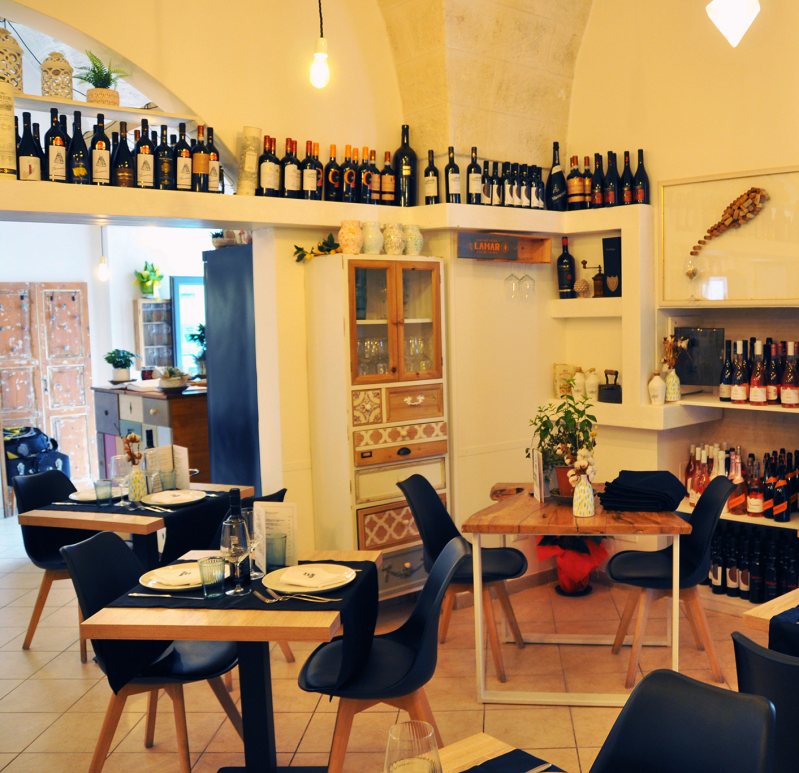 Accogliente interno di un caffè con sedie nere, tavolini, bottiglie di vino sugli scaffali e una calda illuminazione gialla.