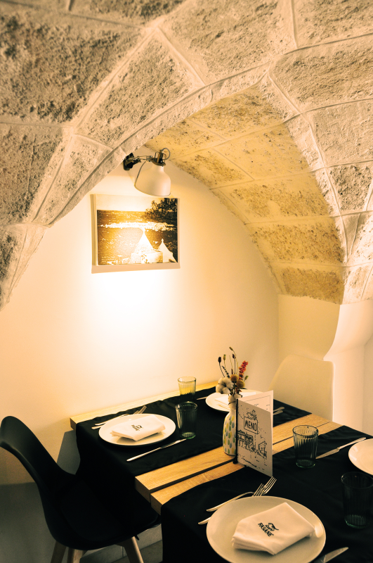 Accogliente angolo pranzo con arco in pietra, tavolo apparecchiato, sedie nere, luci calde e quadri appesi alle pareti.