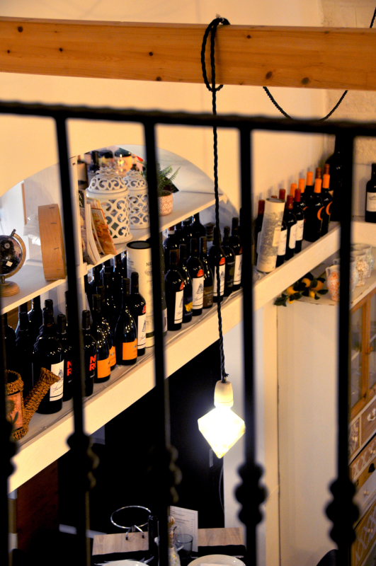 Scaffali di bottiglie di vino in un negozio illuminato da una luce calda, visti attraverso sbarre di ringhiera nere.