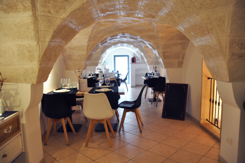 Sala da pranzo con soffitto a volta in pietra, tavoli, sedie e una luminosa finestra in fondo.