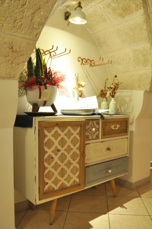 Mobiletto decorativo con fiori e lampada sotto una nicchia ad arco in un corridoio piastrellato.