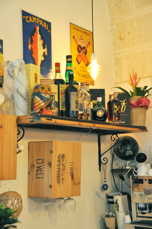Accogliente mensola da cucina con bottiglie, bicchieri, piante e poster vintage su una parete piastrellata.