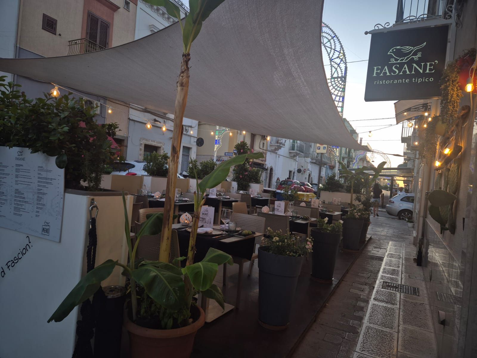Tavolini all'aperto di un caffè lungo una stradina stretta, con tende parasole bianche e un'insegna con la scritta