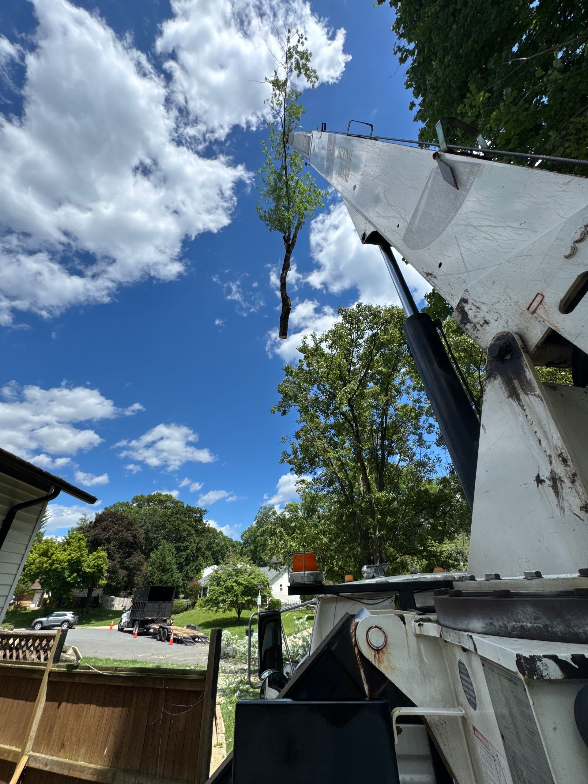Crane arm lifting a white metal structure beside a house under a bright blue sky with clouds
