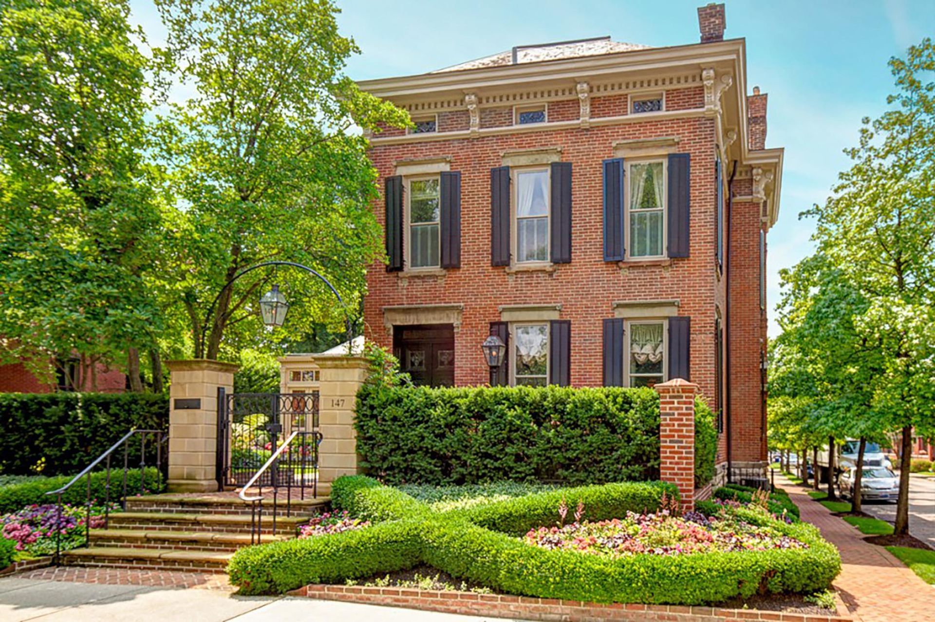 A large brick house with black shutters is surrounded by trees and bushes.