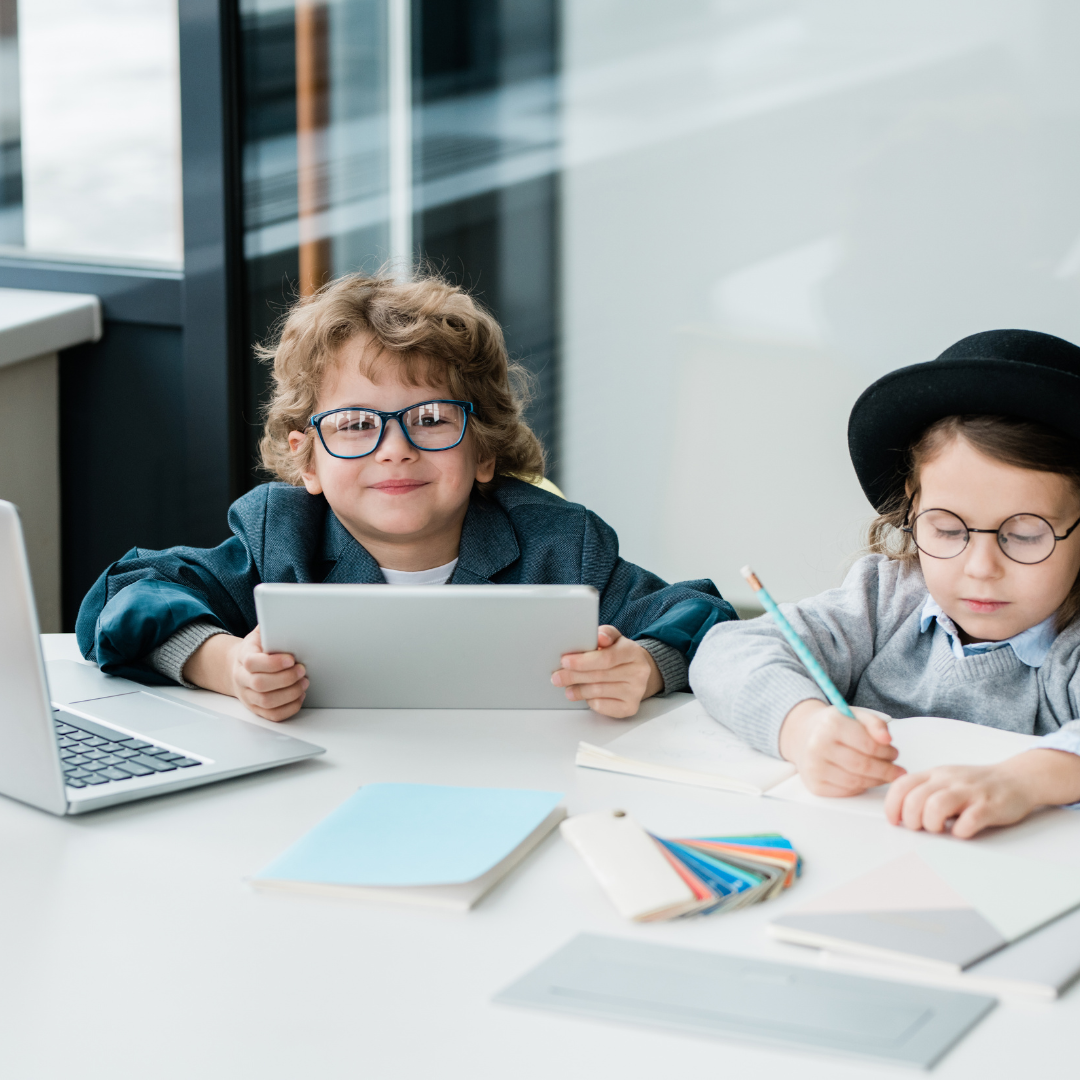 Two children working at a desk with a tablet, laptop, and school supplies in a bright classroom