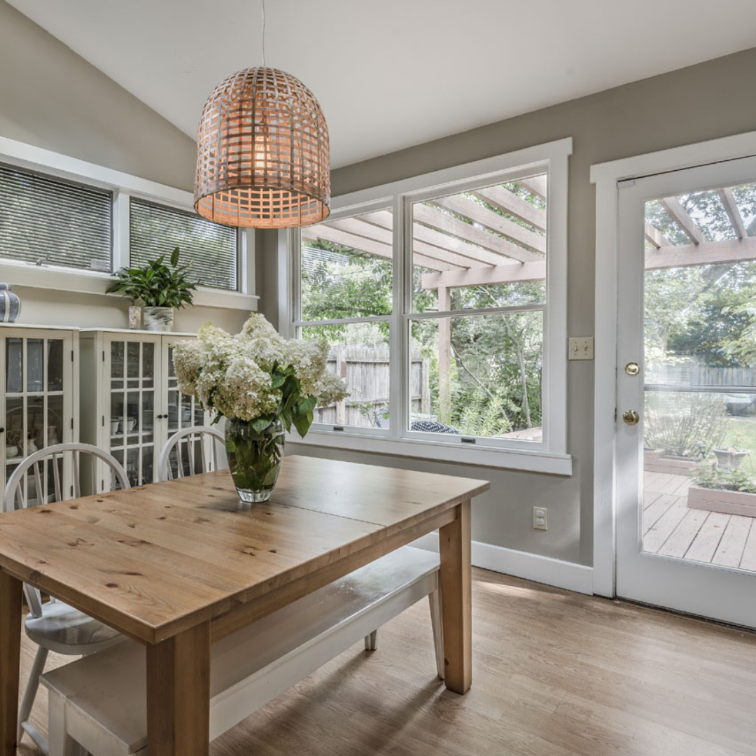 Bright dining nook with wood table, wicker pendant light, and large windows overlooking greenery