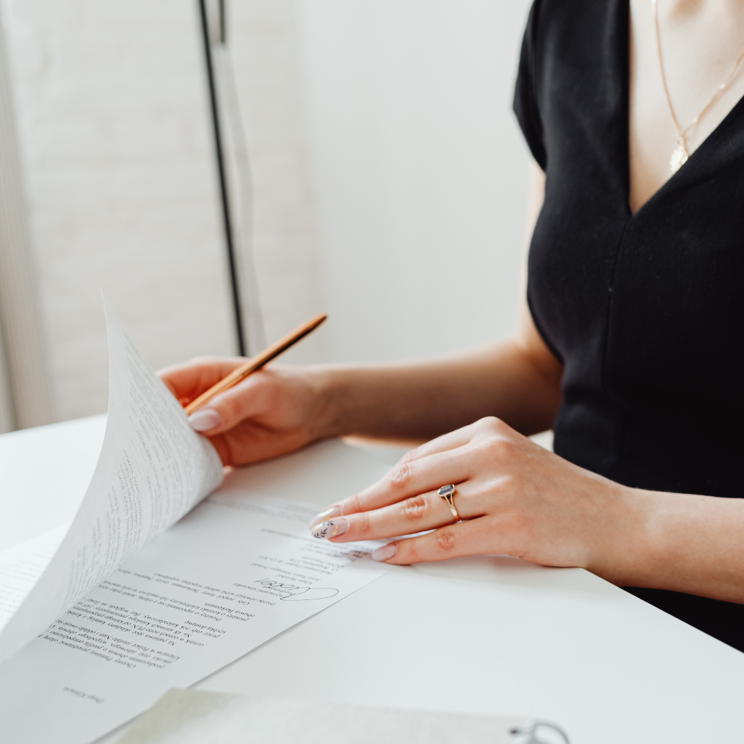 Person writing on a document at a white desk, wearing a black top, with a pen in hand.