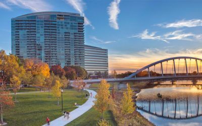 A bridge over a river in a park with a large building in the background.