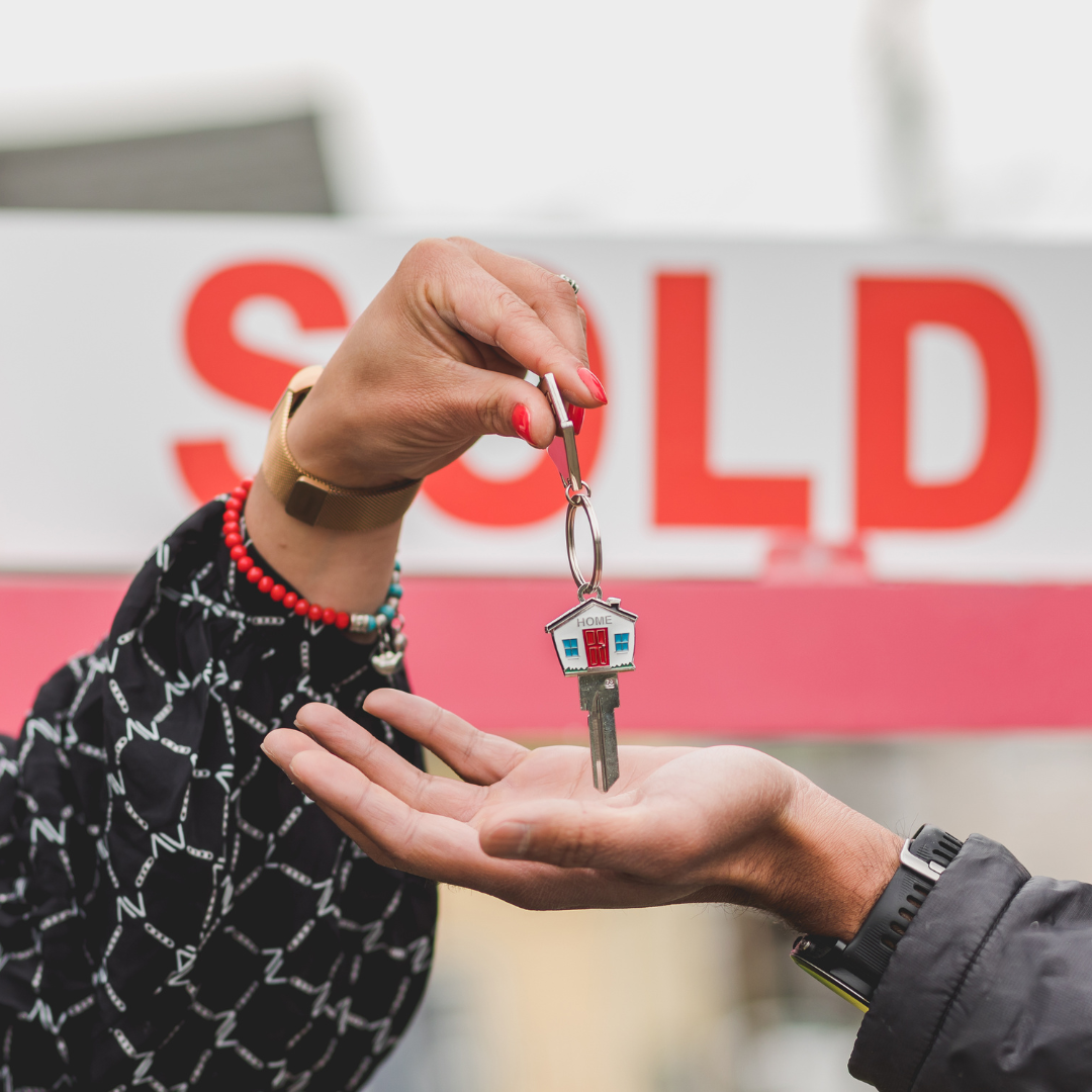 Keys being handed over in front of a red “SOLD” sign.