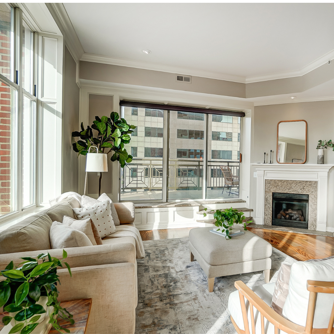 Bright living room with beige sofa, large windows, fireplace, coffee table, and potted plants