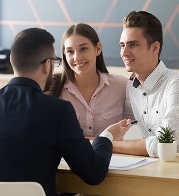 Man in suit talking to smiling couple at a desk; office setting.
