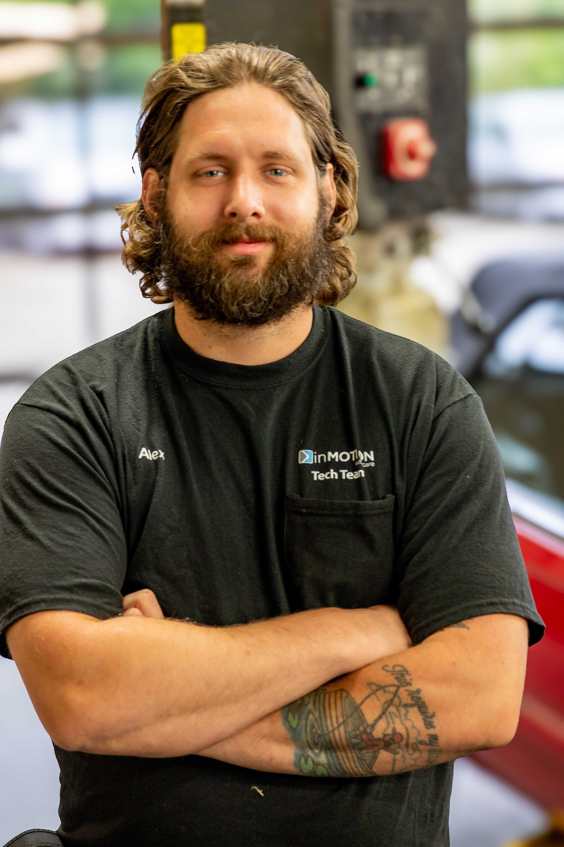 Man standing with arms crossed in a shop, wearing a black T-shirt and sporting a beard and curly hair.