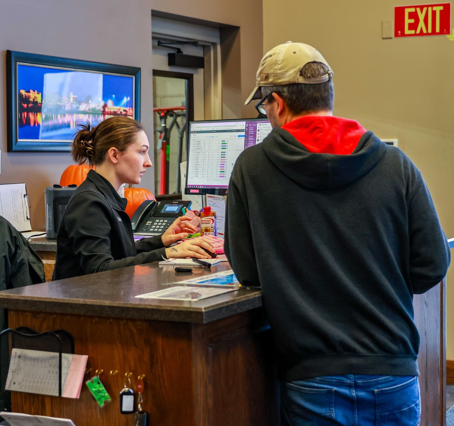 Person at a front desk speaking with a clerk in a lobby, with computer monitors and an exit sign visible.