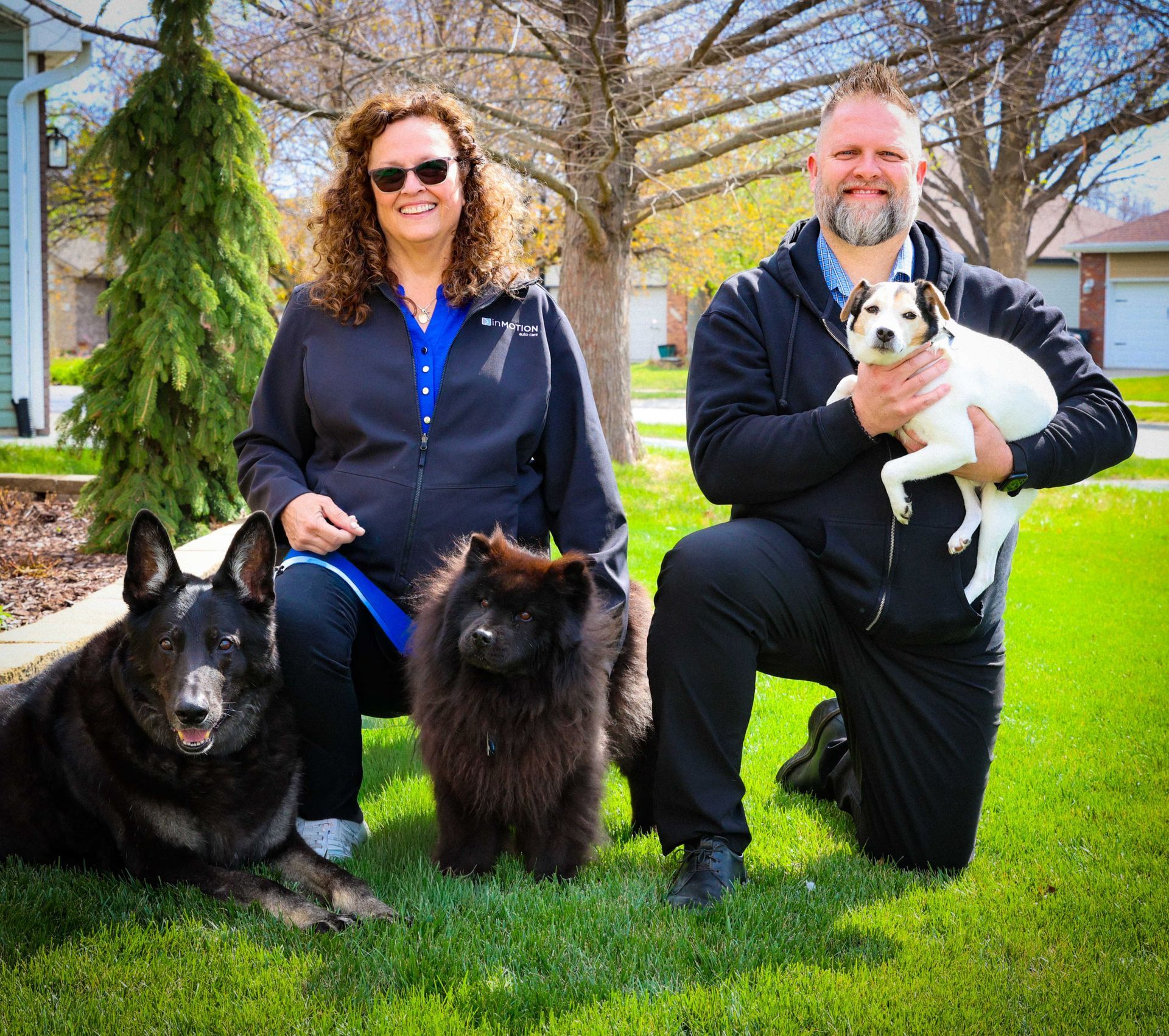 Three people kneel on grass with three dogs in a sunny park.