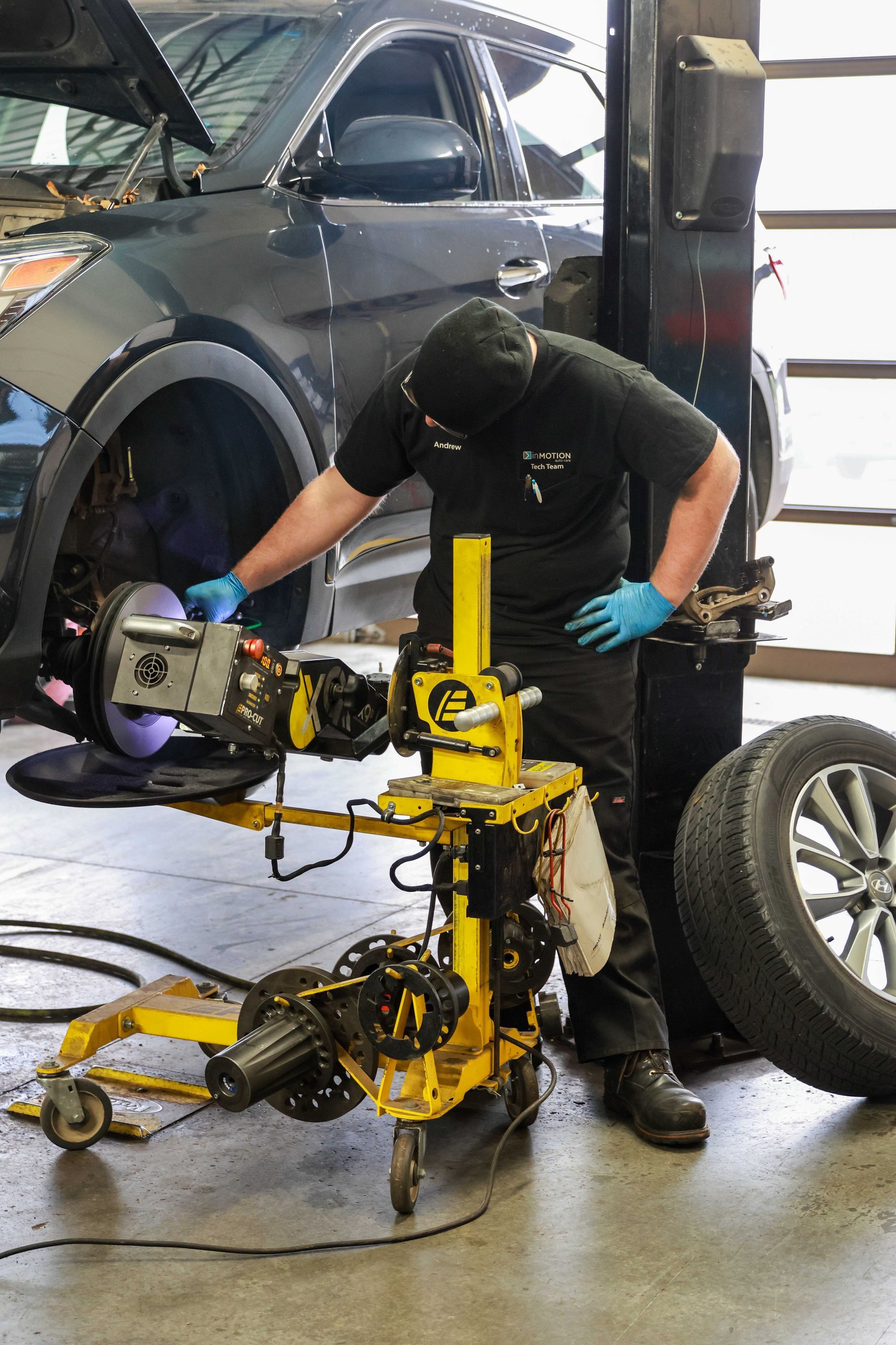 Mechanic in black workwear and blue gloves adjusting a yellow wheel-alignment machine beside a car in a garage