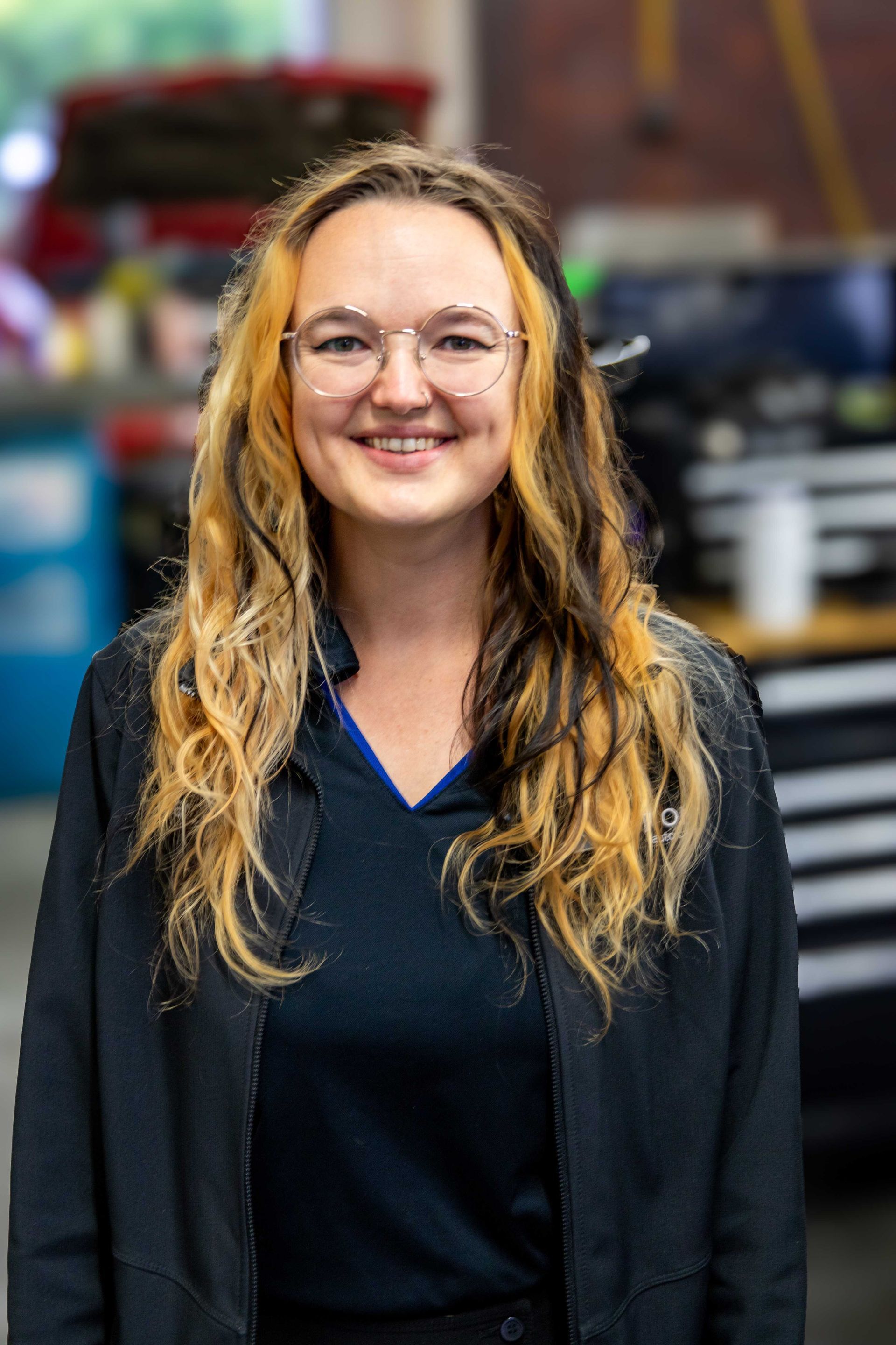 Smiling person with teal hair and glasses wearing a blue and black polo indoors
