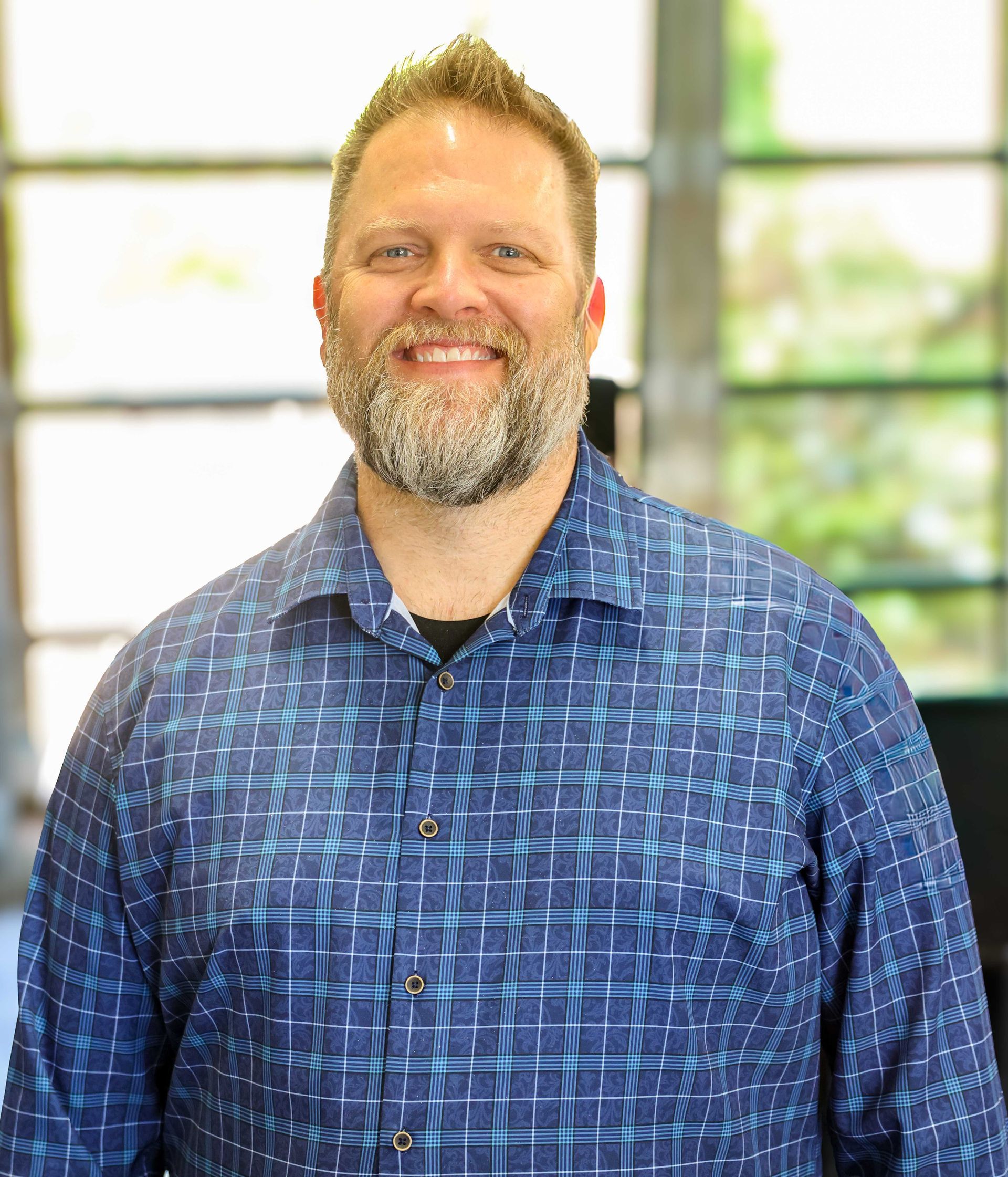 Smiling man in a blue patterned shirt standing indoors by a bright window